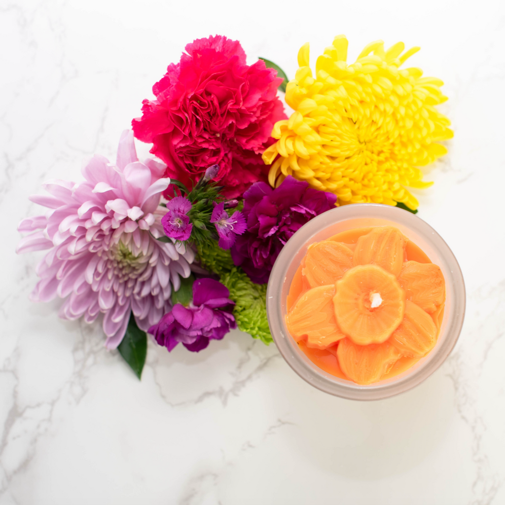 A scented candle with a daffodil flower top design, surrounded by various colorful flowers in a marble background.